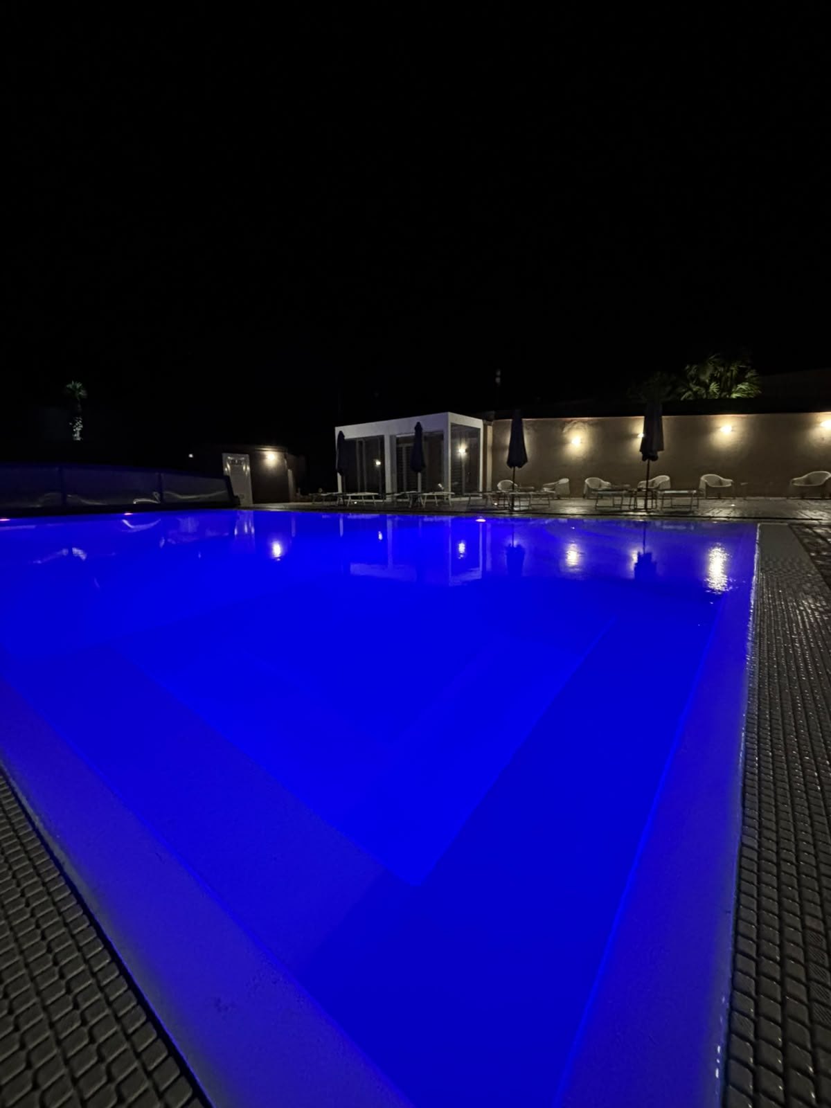 Piscina di Villa Vittoria Relais illuminata di blu di notte, con bordo grigliato, ombrelloni chiusi e poltroncine bianche