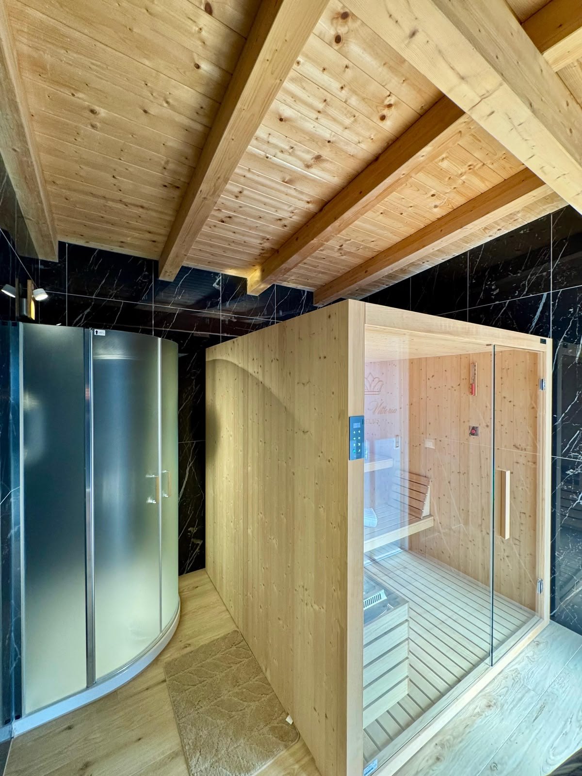 Sauna in legno chiaro con anta in vetro accanto a doccia curva, pareti in marmo nero e soffitto a travi di Villa Vittoria Relais